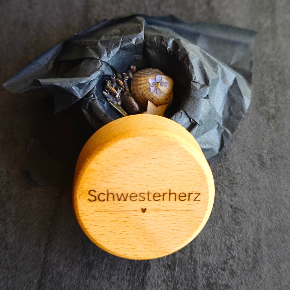 Geöffnete MEMENTOVIA-Box "Ewige Spuren" mit Vergissmeinnicht, Tanne, Stern, Muschel und Amethyst in schwarzem Seidenpapier – symbolische Beigaben für ewige Erinnerung, Schutz und emotionale Heilung. Im Vordergrund der Deckel der Box mit kleiner Herzgravur und dem Wort "Schwesterherz".